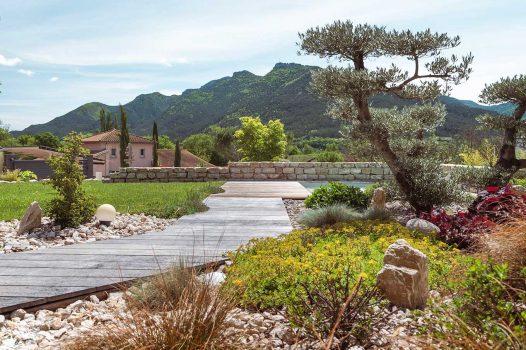 Création d’un espace bien-être extérieur avec l’installation d’un spa encastré à hauteur du sol, harmonieusement intégré dans un jardin paysager. Le spa est bordé par une terrasse en lames de bambou posée sur une structure métallique Terrassteel, assurant durabilité, résistance à l’humidité et finition soignée. Un mur en pierre naturelle vient structurer l’espace et renforcer l’ambiance authentique et chaleureuse. Des massifs paysagers complètent l’ensemble, avec notamment un olivier majestueux en élément central, apportant une touche méditerranéenne à la composition. Aménagement alliant confort, esthétisme et techniques modernes de construction extérieure. Ce projet illustre le savoir-faire d’un paysagiste et pisciniste en région Rhône-Alpes, intervenant à Valence, dans la Drôme et en Ardèche, pour des réalisations harmonieuses et techniques mêlant confort, tradition et modernité.
